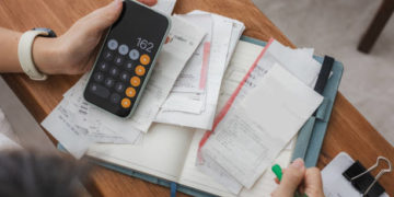 Close up a woman managing her home finances with a smartphone, she reviews financial bills. Concepts include financial planning, taxes, spending, budgeting, and financial challenges.