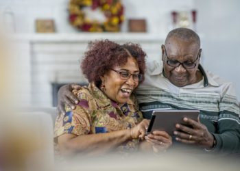 A senior man and woman of African descent are sitting on a couch in their living room. They are having fun watching a video on a tablet computer together.