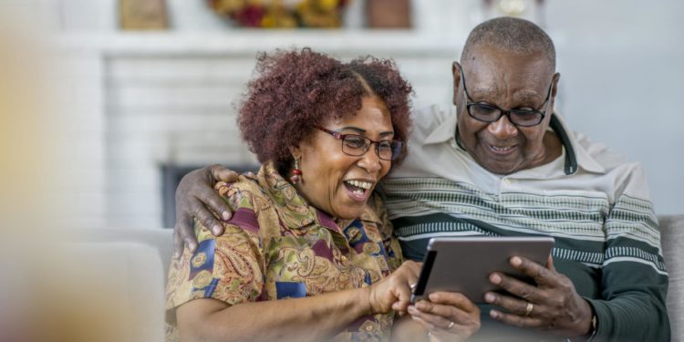 A senior man and woman of African descent are sitting on a couch in their living room. They are having fun watching a video on a tablet computer together.